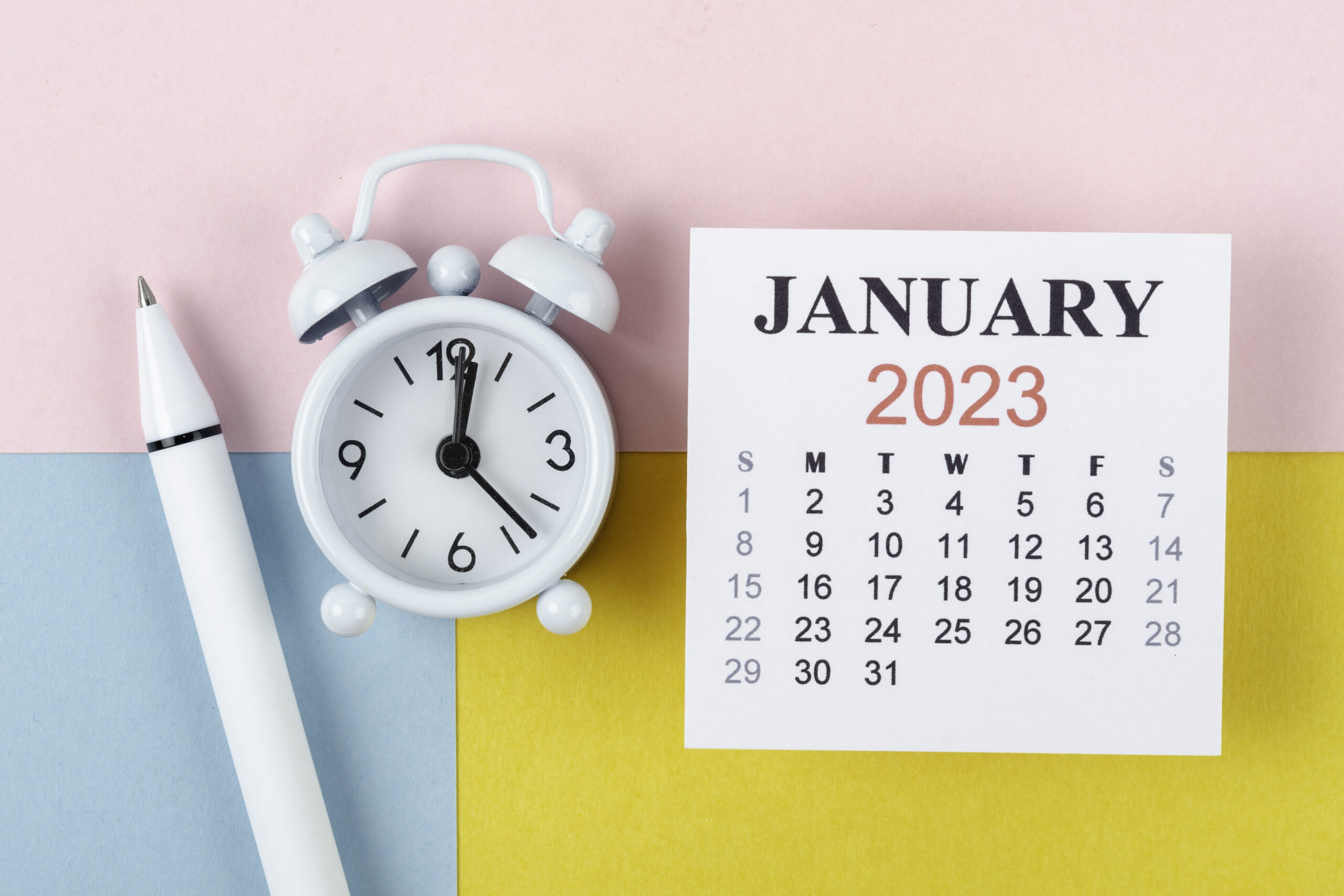 Calendar Desk 2023: January is the month for the organizer to plan and deadline with an alarm clock and white pen on a two-tone paper background.