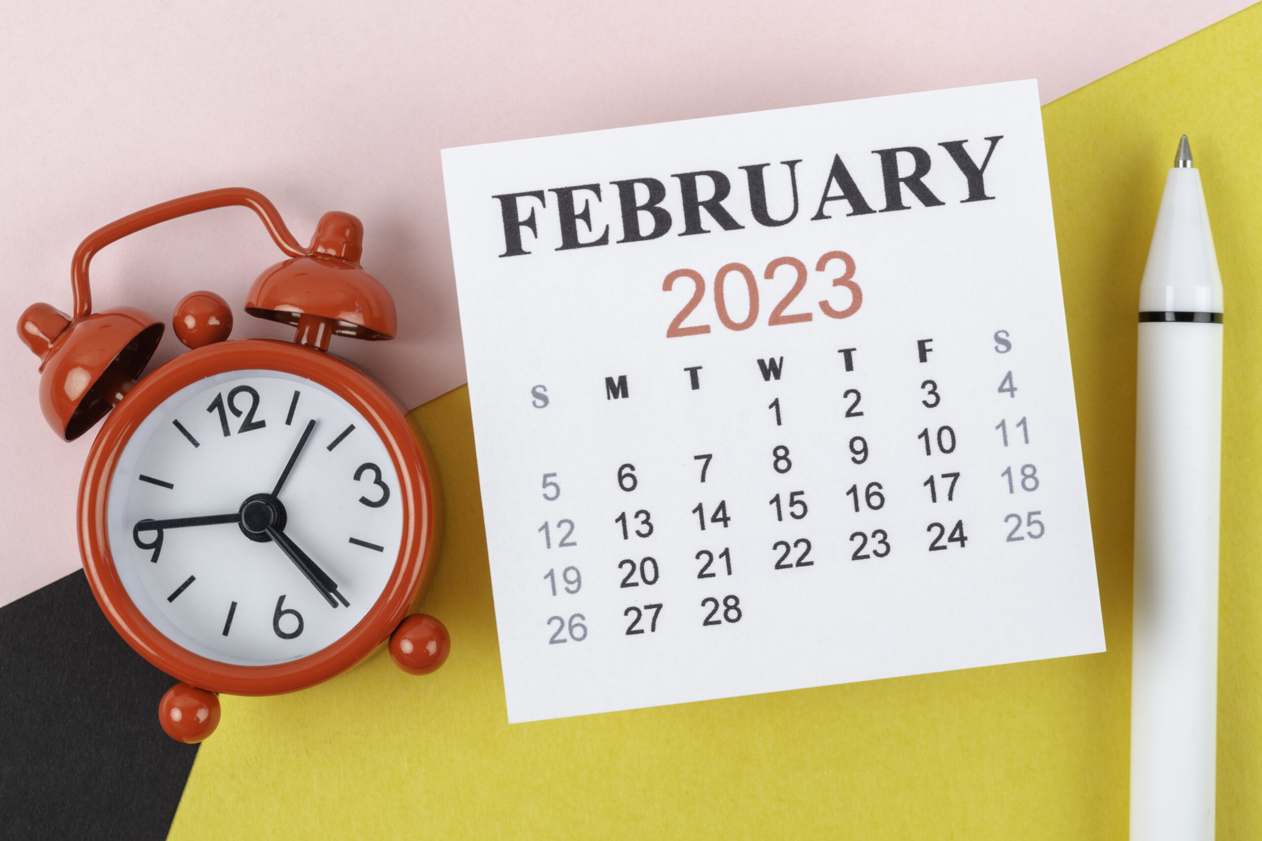 Calendar Desk 2023: February is the month for the organizer to plan and deadline with an alarm clock and white pen on a two-tone paper background.